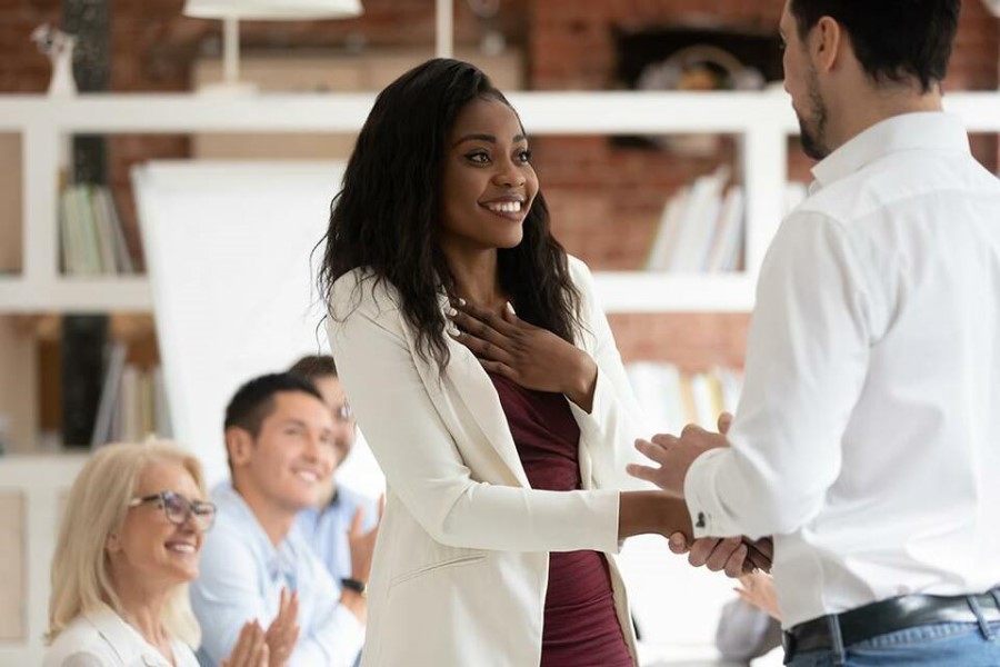 A young Black businesswoman smiles and holds her left hand over her heart while her supervisor shakes her hand. Her coworkers are seen applauding in the background.