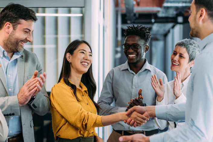 A woman smiles and shakes hands with a manager while he coworkers smile and clap.