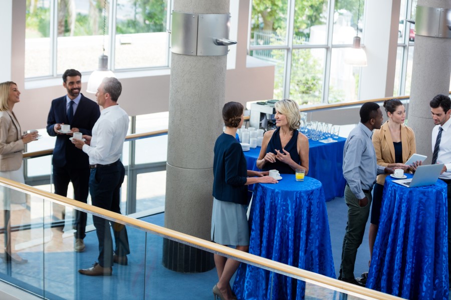 A far away shot of people in business attire mingling around blue cocktail tables in a convention building