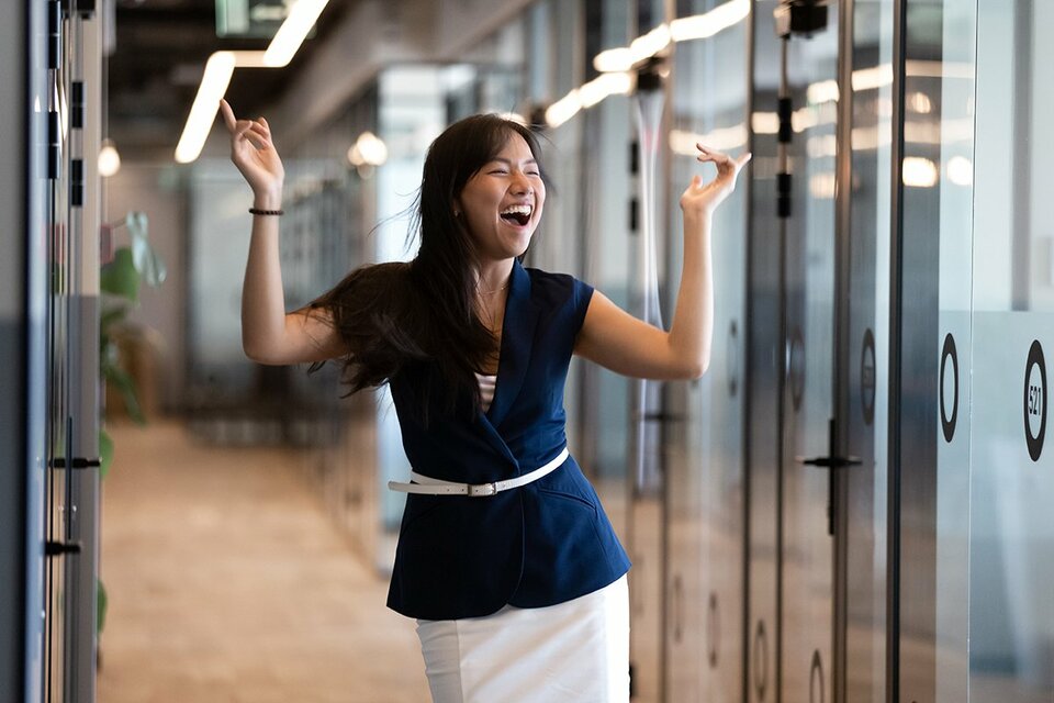 A young businesswoman puts her hands up and smiles as she happily walks through a hallway
